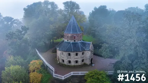 Luchtfoto van een stenen kapel met leien spitsdak in het Valkhof gebied in Nijmegen, omringd door herfstbomen in de mist