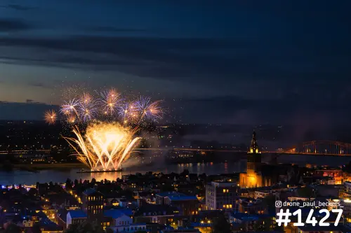 Luchtfoto van vuurwerk boven de Waal in Nijmegen in de avondschemering, met de verlichte Stevenskerk en de Waalbrug