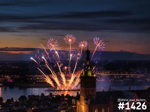 Luchtfoto van vuurwerk boven de Waal in Nijmegen bij schemering, met de toren van de Stevenskerk op de voorgrond