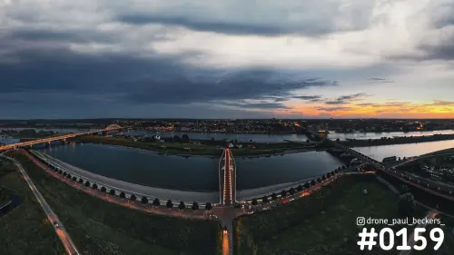 Luchtfoto van de Waal bij Nijmegen-Lent in de avondschemering, met verlichte brug over de nevengeul en oranje gloed