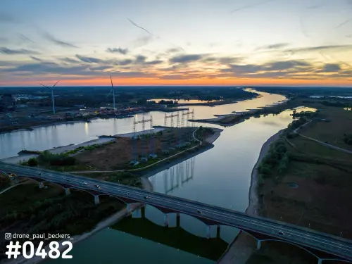Luchtfoto van de Waal en de nevengeul bij Nijmegen in de schemering, met brug, hoogspanningsmasten en windmolens