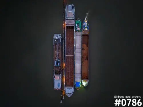 Luchtfoto van vier binnenvaartschepen naast elkaar aan een steiger op de Waal bij Nijmegen, in het donker met verlichte dekken