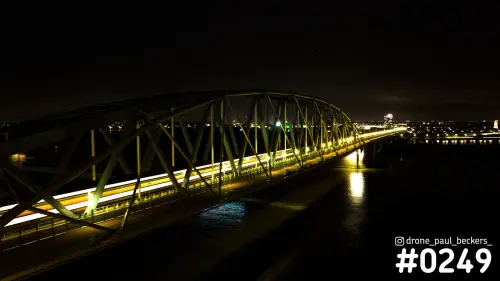Luchtfoto van een stalen boogbrug over de Waal bij Nijmegen in de nacht, met lange lichtstrepen langs het brugdek