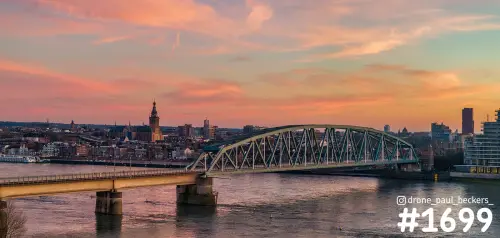 Luchtfoto van de Waal in Nijmegen met een stalen boogbrug en de Stevenskerk onder een roze en oranje avondlucht