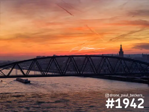 Luchtfoto van stalen boogbrug over de Waal in Nijmegen bij oranje schemerlicht, met Stevenskerk en vrachtschip