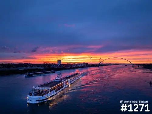 Luchtfoto van een riviercruiseschip op de Waal in Nijmegen bij zonsondergang, met De Oversteek op de achtergrond