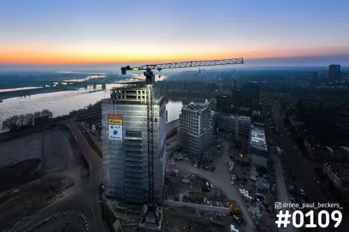 Luchtfoto van een hoogbouw in aanbouw met bouwkraan aan de Waal in Nijmegen, oranje schemering boven de stad en rivier