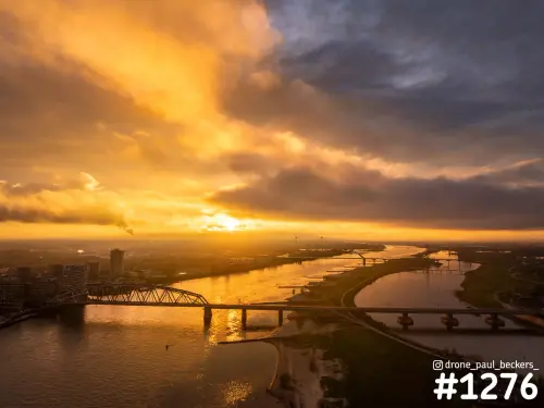 Luchtfoto van de Waal bij Nijmegen met de Oversteek en de spoorbrug in avondlicht, zon onder dramatische wolken