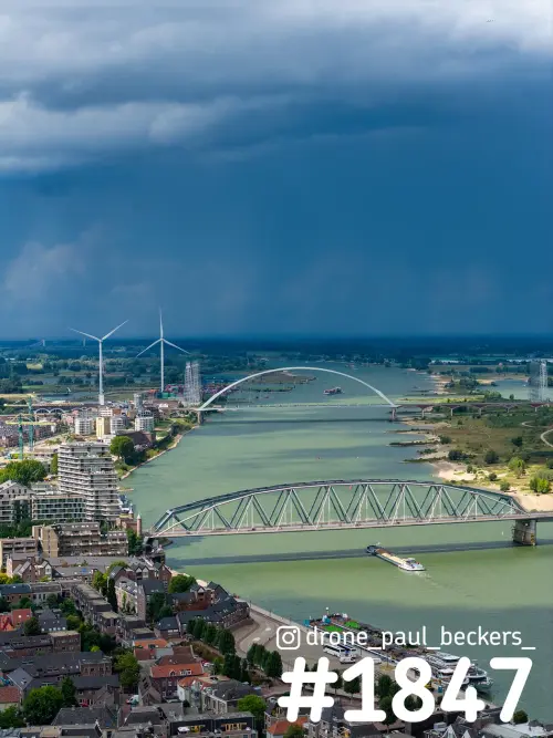 Luchtfoto van de Waal bij Nijmegen met De Oversteek en een stalen vakwerkbrug onder een donkere regenbui