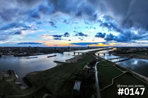 Luchtfoto van de Waal bij Nijmegen in de schemering, met De Oversteek en een oranje gloed onder blauwe wolken