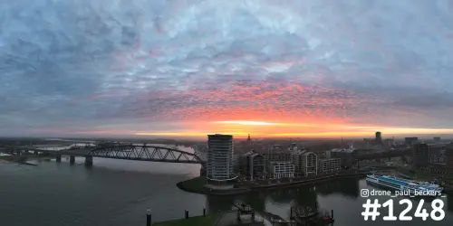 Panorama van de Waal in Nijmegen met oranje gloed onder wolkendek, stalen brug links en hoogbouw aan de Handelskade
