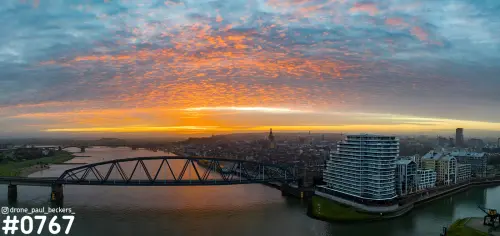 Panorama van Nijmegen aan de Waal met oranje wolkenlucht, een stalen brug, de Nimbus en de toren van de Stevenskerk