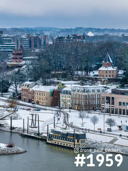 Luchtfoto van de besneeuwde oever van de Waal in Nijmegen, met een boot aan de kade en het Kronenburgerpark erachter