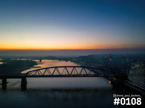 Luchtfoto van een stalen brug over de Waal in Nijmegen bij avondgloed, met De Oversteek in de verte