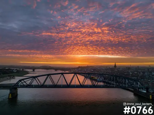 Luchtfoto van stalen brug over de Waal in Nijmegen bij zonsopkomst, met rode wolkenlucht en Stevenskerk aan de horizon