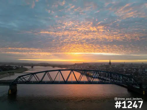 Luchtfoto van een stalen brug over de Waal in Nijmegen bij zonsopkomst, met oranje schapenwolken en de Stevenskerk