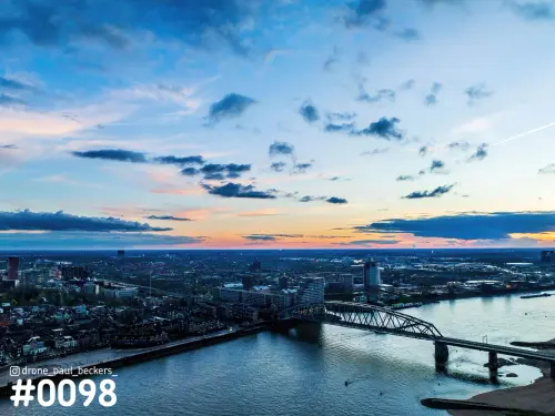 Luchtfoto van de Waal en de Waalkade in Nijmegen in de schemering, met een stalen boogbrug en een blauwe wolkenlucht