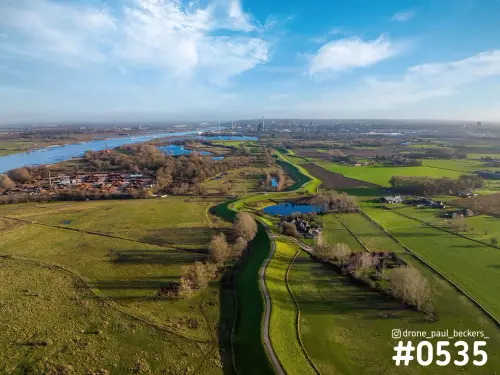 Luchtfoto van de Waal bij Weurt met groene dijk, uiterwaarden en polderland onder blauwe lucht met wolkenslierten