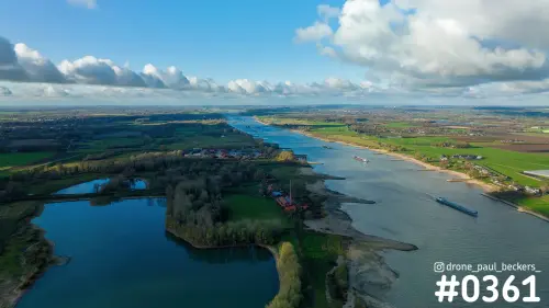 Luchtfoto van de Waal bij Weurt met vrachtschepen, een plas en zandige oevers onder blauwe lucht met stapelwolken