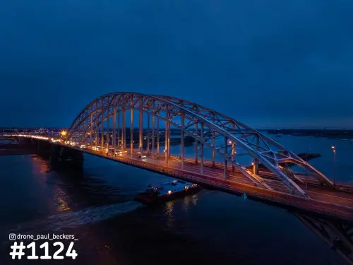 Luchtfoto van de Waalbrug in Nijmegen in het blauwe uur, met verlichte boogconstructie en een vrachtschip op de Waal