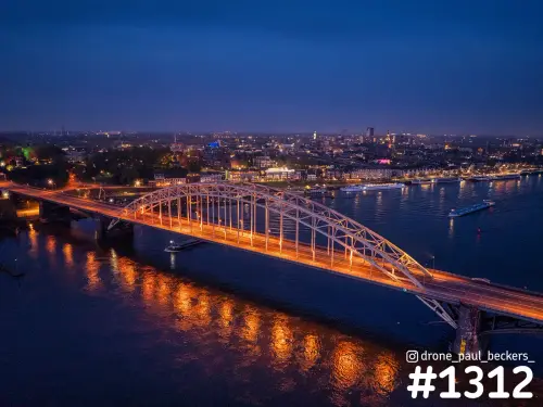 Luchtfoto van de Waalbrug in Nijmegen in het blauwe uur, met verlichte boog en oranje weerspiegeling in de Waal