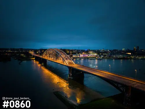 Luchtfoto van de verlichte Waalbrug in Nijmegen na zonsondergang, met gouden weerspiegeling in de Waal