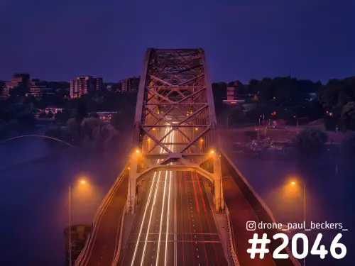 Luchtfoto van de Waalbrug in Nijmegen in de avondschemering, met lichtsporen van verkeer en mist boven de Waal