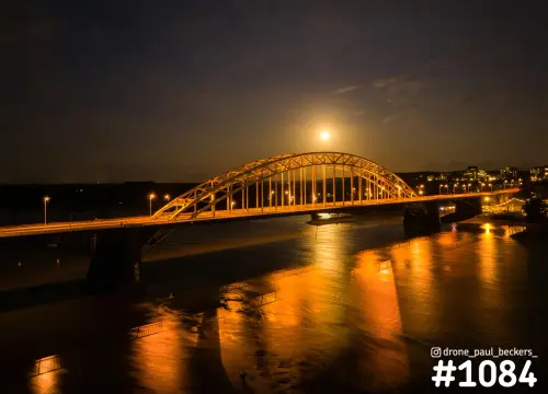Luchtfoto van de verlichte Waalbrug in Nijmegen 's avonds, met de maan boven de boog en gouden weerspiegeling in de Waal