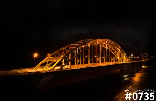 Verlichte boogbrug van de Waalbrug in Nijmegen in het donker, met oranje licht en weerspiegeling in de Waal