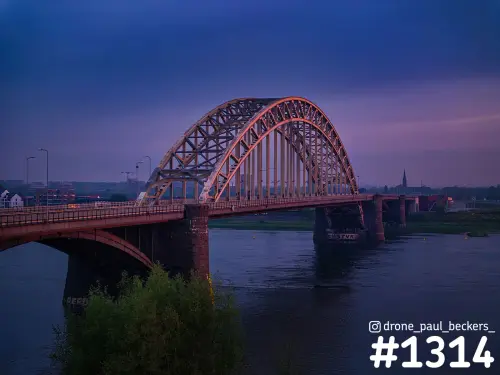 Luchtfoto van de Waalbrug in Nijmegen na zonsondergang, met de warm verlichte boog boven de Waal en een paarse lucht