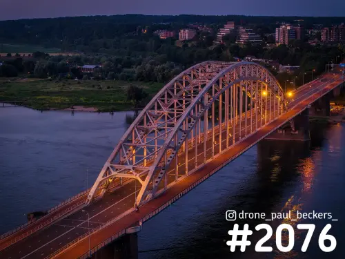 Luchtfoto van de Waalbrug in Nijmegen in de schemering, met verlichte boogconstructie en lantaarns die weerkaatsen in de Waal