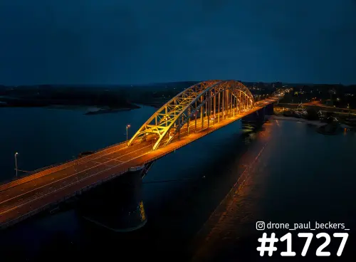 Luchtfoto van de verlichte Waalbrug in Nijmegen in de avondschemering, met oranje licht op de boog en weerspiegeling in de Waal