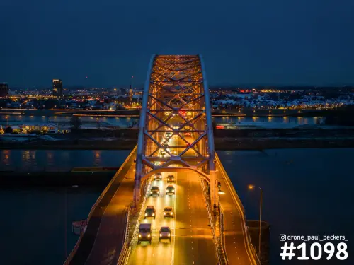 Luchtfoto van de verlichte boog van de Waalbrug in Nijmegen in de avondschemering, met sneeuw op de daken van Lent