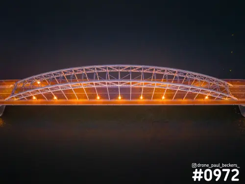 Luchtfoto van de Waalbrug in Nijmegen van recht boven, met verlichte stalen boog en oranje licht boven de donkere Waal