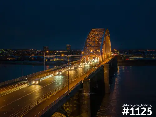 Luchtfoto van de verlichte boogbrug Waalbrug in Nijmegen na zonsondergang, met autolichten en lichtreflecties op de Waal