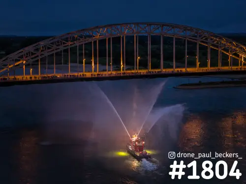 Luchtfoto van de Waalbrug in Nijmegen in de avondschemering, met een boot die waterstralen spuit op de Waal