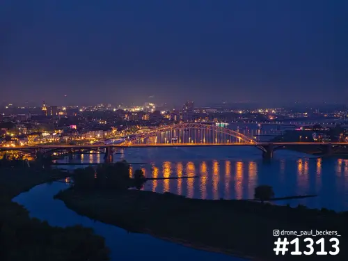 Luchtfoto van de verlichte Waalbrug in Nijmegen in de avondschemering, met oranje lichtreflecties in de Waal en de stad op de achtergrond