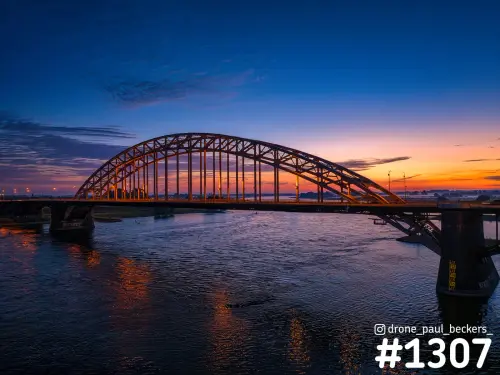 Luchtfoto van de Waalbrug in Nijmegen na zonsondergang, met oranje gloed aan de horizon en weerspiegeling in de Waal
