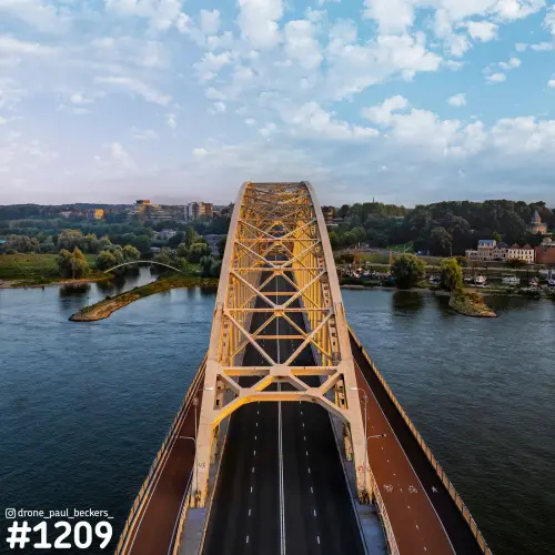 Luchtfoto van de gele boogconstructie van de Waalbrug in Nijmegen in avondlicht, met de rivier de Waal aan weerszijden