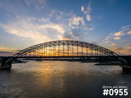 Dronefoto van de Waalbrug in Nijmegen in het avondlicht, met oranje gloed boven de Waal en mensen op het brugdek