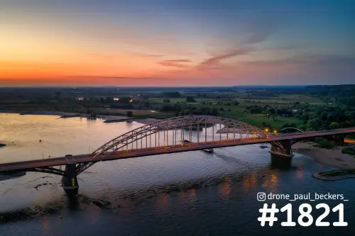 Luchtfoto van de Waalbrug in Nijmegen na zonsondergang, met oranje avondlucht boven de Waal en de Ooijpolder