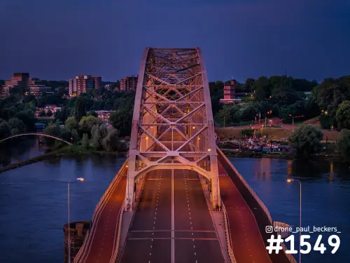 Luchtfoto van de Waalbrug in Nijmegen in de avondschemering, met verlichte stalen boog en lege rijbanen boven de Waal