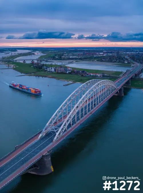 Luchtfoto van de Waalbrug in Nijmegen in de avondschemering, met een containerschip op de Waal en Lent op de achtergrond