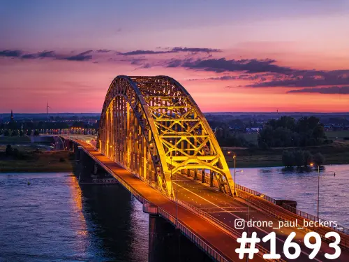 Luchtfoto van de geel verlichte boogbrug Waalbrug in Nijmegen in de avondschemering, met roze lucht boven de Waal