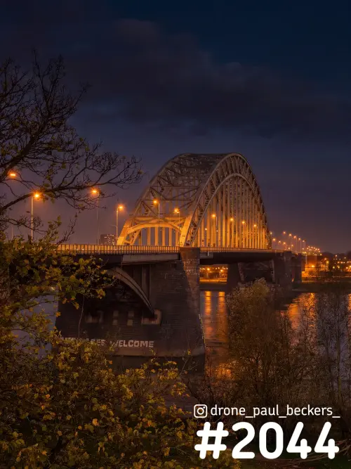 Waalbrug in Nijmegen in de avondschemering, met verlichte boog, oranje lantaarns en weerspiegeling in de Waal