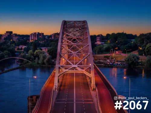 Luchtfoto van de Waalbrug in Nijmegen in de avondschemering, met verlichte stalen boog en een oranje horizon