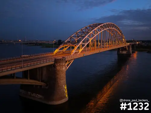 Luchtfoto van de verlichte Waalbrug in Nijmegen in het blauwe uur, met de boogbrug en oranje reflecties op de rivier de Waal