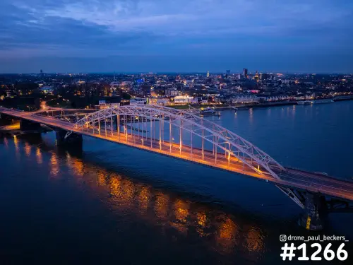 Luchtfoto van de verlichte Waalbrug in Nijmegen in het blauwe uur, met oranje reflecties in de Waal en de stad op de achtergrond