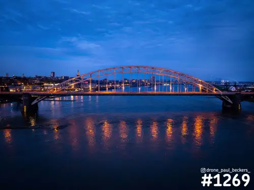 Luchtfoto van de verlichte Waalbrug in Nijmegen in het blauwe uur, met oranje lichtreflecties in de Waal