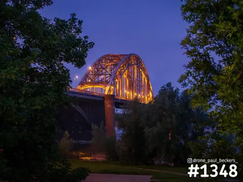 De verlichte Waalbrug in Nijmegen in het blauwe uur, gezien tussen bomen door met oranje licht op de stalen bogen